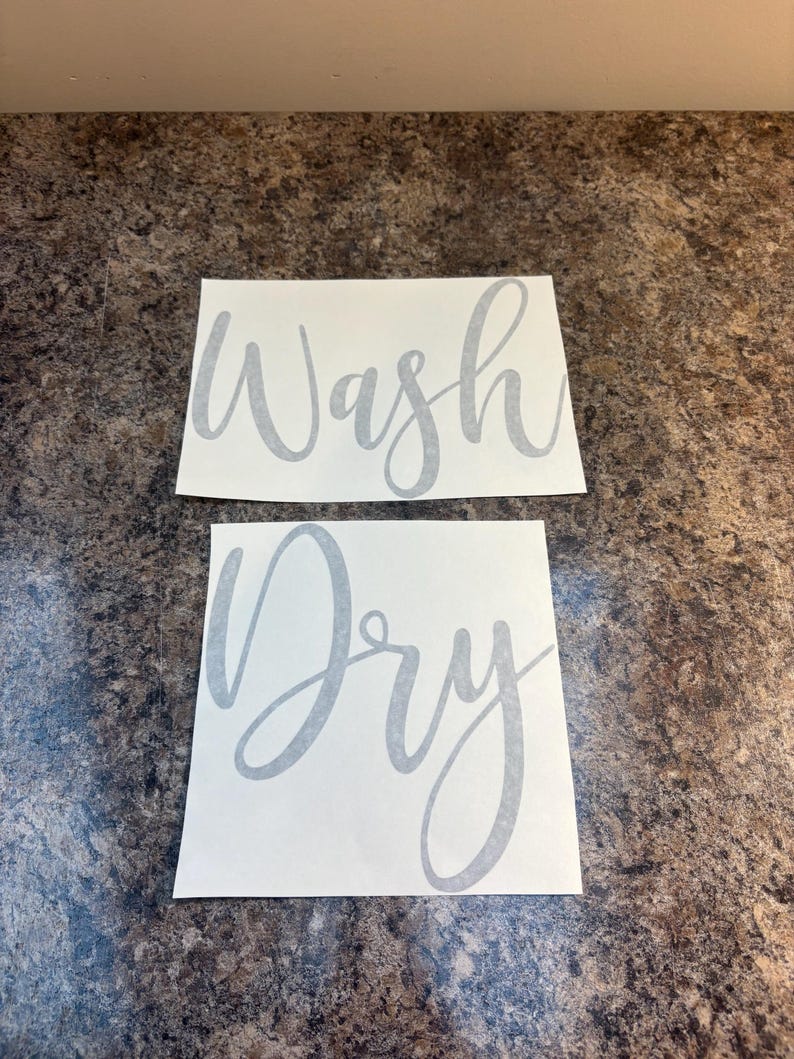 Puede incluir: Dos calcoman&iacute;as rectangulares blancas con texto gris en cursiva. La calcoman&iacute;a superior dice "Wash", y la calcoman&iacute;a inferior dice "Dry". Las calcoman&iacute;as se muestran sobre una superficie marr&oacute;n moteada.