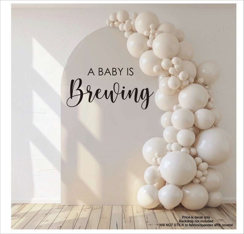 May include: A cream-colored arch with the text "A BABY IS Brewing" in black script. A cascade of white balloons adorns the right side. The image is a decal, not a backdrop, and will not stick to fabrics or spandex arch covers.