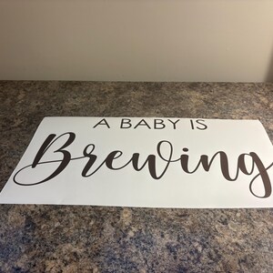 May include: A white rectangular sign with the phrase "A BABY IS Brewing" in a dark brown, elegant script. The sign is placed on a speckled, brown surface.