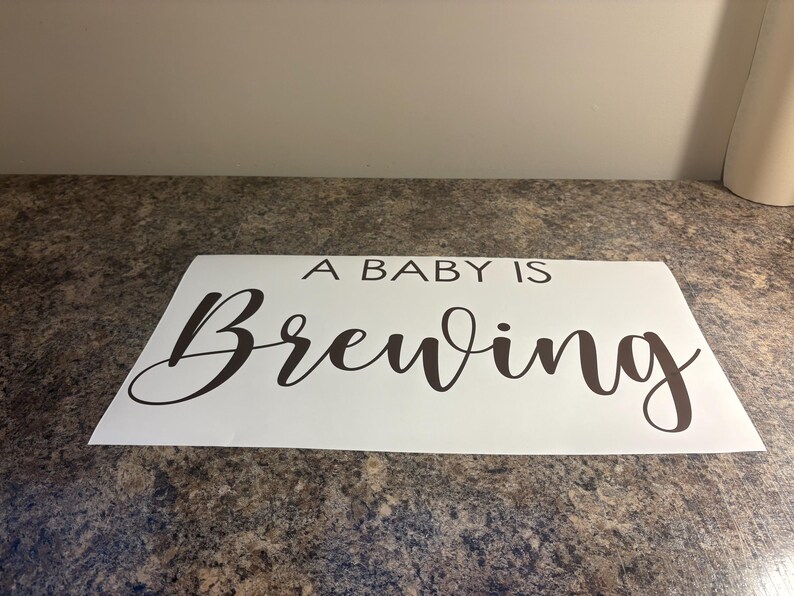 May include: A white rectangular sign with the phrase "A BABY IS Brewing" in a dark brown, elegant script. The sign is placed on a speckled, brown surface.