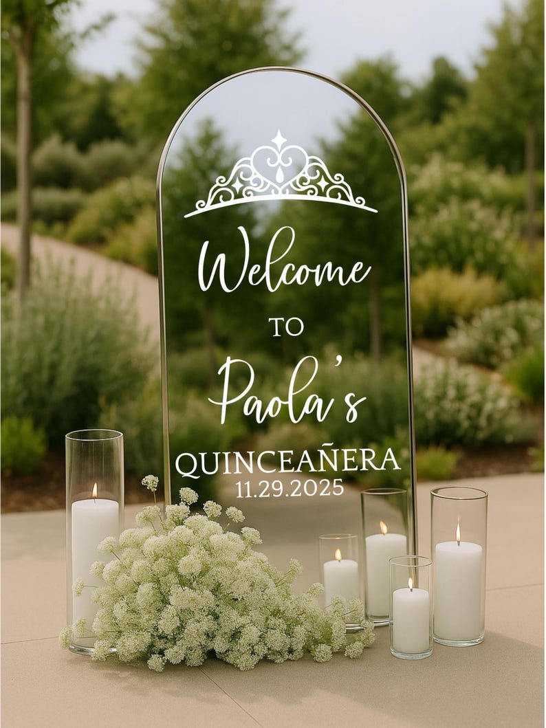 May include: A clear, arched mirror with the words "Welcome to Paola's Quinceañera 11.29.2025" in white script. The mirror is decorated with a crown design. White candles in glass cylinders and white flowers are arranged in front of the mirror.