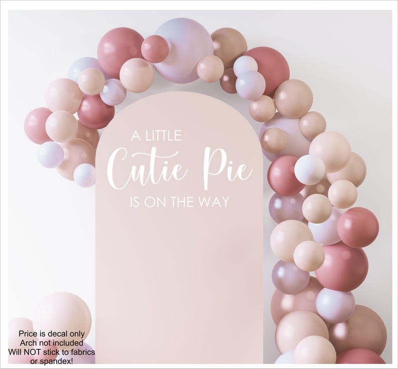 May include: A light pink arched backdrop decal featuring the white text A LITTLE Cutie Pie IS ON THE WAY, surrounded by a decorative balloon garland in various shades of dusty rose, mauve, beige, and pale lavender, arranged in an arch shape.
