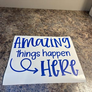 Puede incluir: Una calcoman&iacute;a de vinilo blanca cuadrada con el texto "Amazing things happen HERE" en una fuente azul, negrita y estilo manuscrito. La palabra "Amazing" est&aacute; en la l&iacute;nea superior, "things happen" en el medio y "HERE" en la parte inferior con una flecha azul.