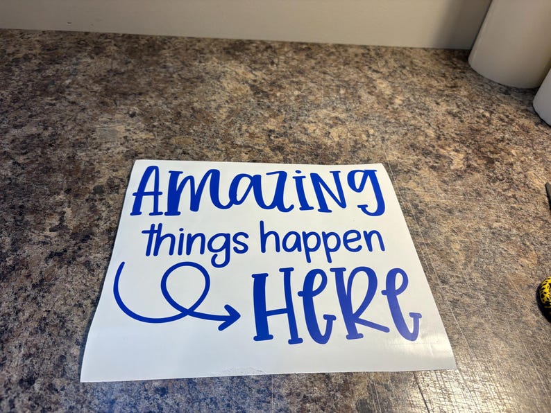 Puede incluir: Una calcoman&iacute;a de vinilo blanca cuadrada con el texto "Amazing things happen HERE" en una fuente azul, negrita y estilo manuscrito. La palabra "Amazing" est&aacute; en la l&iacute;nea superior, "things happen" en el medio y "HERE" en la parte inferior con una flecha azul.