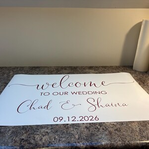 Może przedstawiać: Biała prostokątna naklejka winylowa z ciemnoczerwonym tekstem o treści welcome TO OUR WEDDING Chad & Shaina 09.12.2026. Projekt wykorzystuje połączenie eleganckiej czcionki kaligraficznej oraz prostego kroju pisma bezszeryfowego.