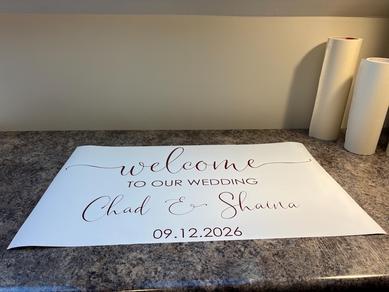 Może przedstawiać: Biała prostokątna naklejka winylowa z ciemnoczerwonym tekstem o treści welcome TO OUR WEDDING Chad & Shaina 09.12.2026. Projekt wykorzystuje połączenie eleganckiej czcionki kaligraficznej oraz prostego kroju pisma bezszeryfowego.