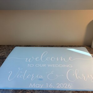 Może przedstawiać: Jasnoniebieski prostokątny znak z białym napisem "welcome TO OUR WEDDING Victoria & Chris May 16, 2026". Tekst wykorzystuje połączenie eleganckiego pisma odręcznego oraz wyraźnych, wielkich liter bezszeryfowych dla daty i szczeg&oacute;ł&oacute;w wydarzenia.