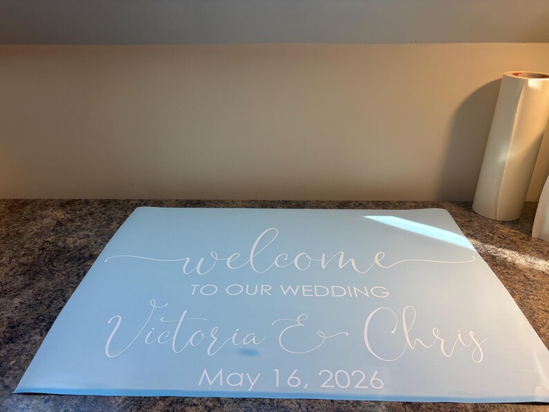 Może przedstawiać: Jasnoniebieski prostokątny znak z białym napisem "welcome TO OUR WEDDING Victoria & Chris May 16, 2026". Tekst wykorzystuje połączenie eleganckiego pisma odręcznego oraz wyraźnych, wielkich liter bezszeryfowych dla daty i szczeg&oacute;ł&oacute;w wydarzenia.