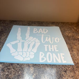 May include: A light blue square decal featuring a white graphic of a skeleton hand making a peace sign. The text "BAD" is printed in a bold sans-serif font, followed by the word "two" in a cursive script, and "THE BONE" in a bold sans-serif font.