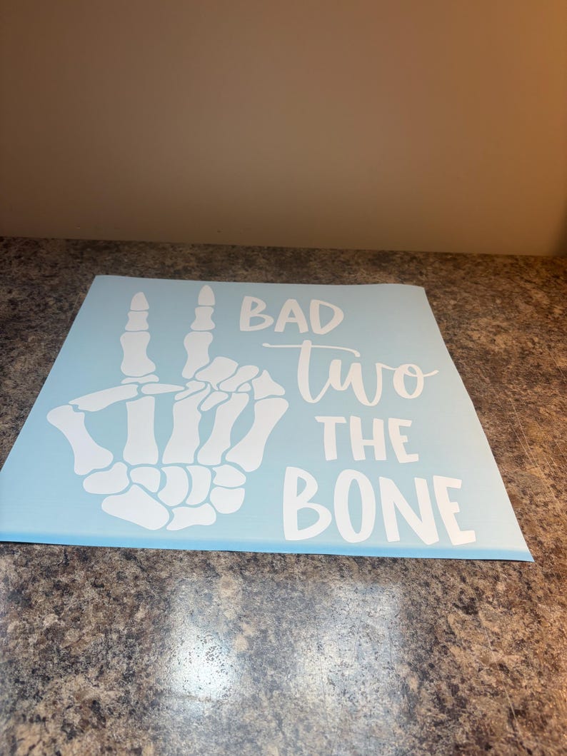 May include: A light blue square decal featuring a white graphic of a skeleton hand making a peace sign. The text "BAD" is printed in a bold sans-serif font, followed by the word "two" in a cursive script, and "THE BONE" in a bold sans-serif font.
