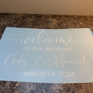 Może przedstawiać: Jasnoniebieski prostokątny znak z białym napisem welcome TO OUR WEDDING Cody & Hannah March 14, 2026. Słowo welcome oraz imiona Cody i Hannah są napisane kursywą, podczas gdy pozostały tekst jest wykonany czcionką bezszeryfową.