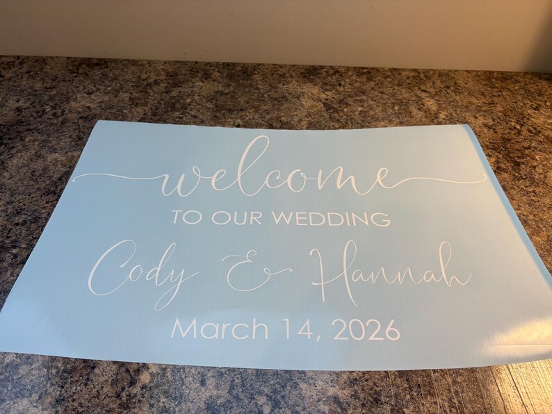 Może przedstawiać: Jasnoniebieski prostokątny znak z białym napisem welcome TO OUR WEDDING Cody & Hannah March 14, 2026. Słowo welcome oraz imiona Cody i Hannah są napisane kursywą, podczas gdy pozostały tekst jest wykonany czcionką bezszeryfową.