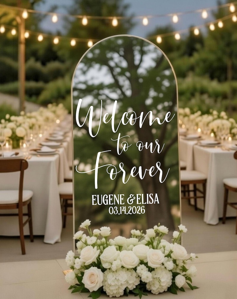May include: An arched mirror wedding welcome sign with a gold frame, featuring white script text that reads Welcome to our Forever, followed by EUGENE & ELISA and the date 03.14.2026, decorated with a base arrangement of white roses and hydrangeas.