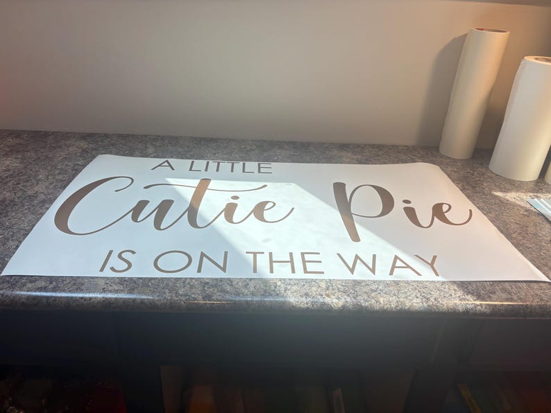 May include: A rectangular white vinyl decal featuring the brown text A LITTLE Cutie Pie IS ON THE WAY. The words A LITTLE and IS ON THE WAY are in a simple sans-serif font, while the words Cutie Pie are in a larger, elegant script font.