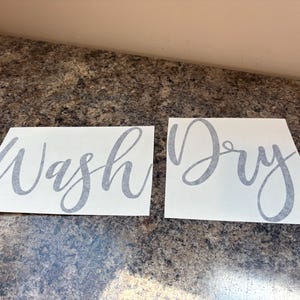 Puede incluir: Dos calcoman&iacute;as rectangulares blancas con texto en cursiva gris. Una dice "Wash" y la otra "Dry". Las calcoman&iacute;as est&aacute;n sobre una superficie marr&oacute;n moteada.
