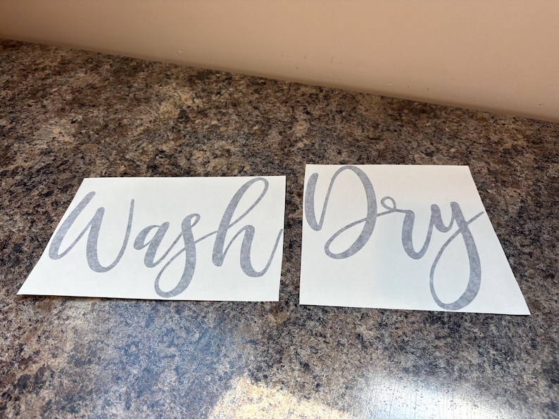 Puede incluir: Dos calcoman&iacute;as rectangulares blancas con texto en cursiva gris. Una dice "Wash" y la otra "Dry". Las calcoman&iacute;as est&aacute;n sobre una superficie marr&oacute;n moteada.