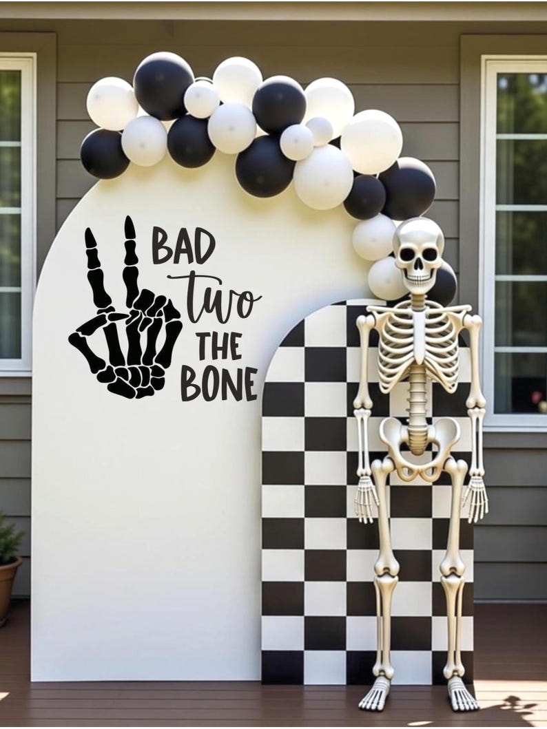 May include: A white arched backdrop with a black skeleton hand making a peace sign and the text "BAD two THE BONE" in black. A smaller checkered arch stands beside it, topped with a black and white balloon garland and a full-body skeleton figure.