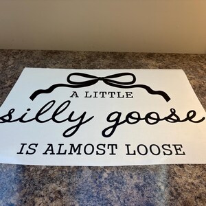 May include: White rectangular sign with black text that reads "A LITTLE silly goose IS ALMOST LOOSE". The text is in a mix of cursive and block letters, with a decorative bow above the word "A LITTLE".