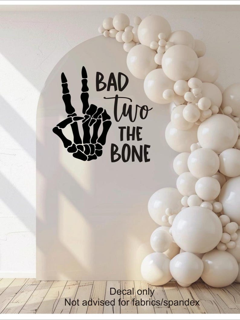 May include: A black vinyl wall decal featuring a skeleton hand making a peace sign gesture, accompanied by the text "BAD Two THE BONE" in a mix of bold and script fonts. The decal is applied to a smooth, arched surface next to white balloons.