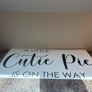 May include: A rectangular white sign featuring black text that reads A LITTLE Cutie Pie IS ON THE WAY. The word Cutie is written in a cursive script font, while the remaining words are displayed in a simple, clean, uppercase sans-serif typeface.