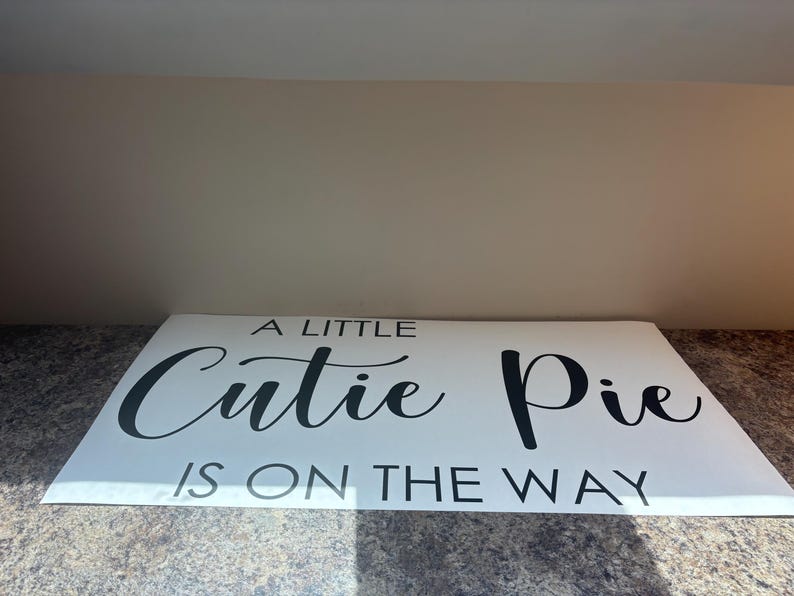 May include: A rectangular white sign featuring black text that reads A LITTLE Cutie Pie IS ON THE WAY. The word Cutie is written in a cursive script font, while the remaining words are displayed in a simple, clean, uppercase sans-serif typeface.