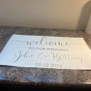Może przedstawiać: Prostokątny biały znak z ciemnym napisem o treści "welcome TO OUR WEDDING John & Brittney 05.02.2026". Tekst łączy płynną czcionkę skryptową dla imion i powitania z wyraźną czcionką bezszeryfową użytą dla daty wydarzenia.