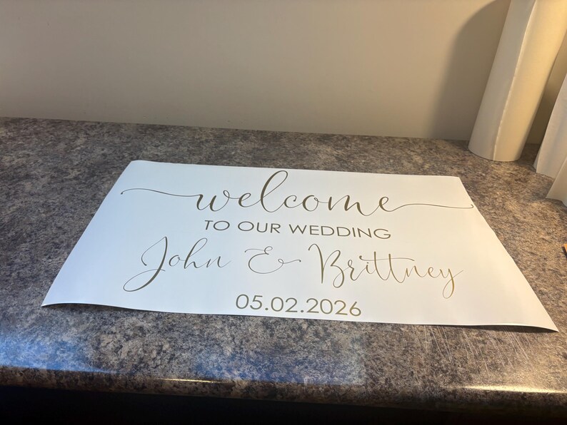Może przedstawiać: Prostokątny biały znak z ciemnym napisem o treści "welcome TO OUR WEDDING John & Brittney 05.02.2026". Tekst łączy płynną czcionkę skryptową dla imion i powitania z wyraźną czcionką bezszeryfową użytą dla daty wydarzenia.