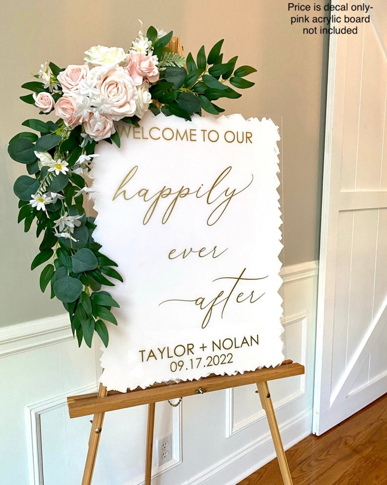 Peut inclure: Une pancarte en acrylique blanche avec des lettres dor&eacute;es qui disent "Bienvenue dans notre bonheur pour toujours Taylor + Nolan 09.17.2022". La pancarte est d&eacute;cor&eacute;e d'une guirlande de fleurs blanches et de feuilles vertes.