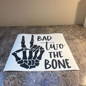 May include: A white square decal featuring a black graphic of a skeleton hand making a peace sign. The text "BAD" is printed in a bold sans-serif font, "two" in a cursive script, and "THE BONE" in a bold sans-serif font, all in black ink.