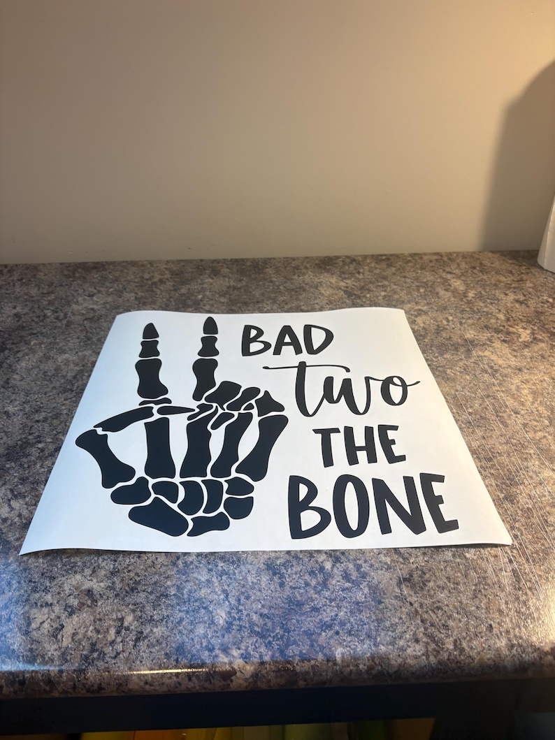 May include: A white square decal featuring a black graphic of a skeleton hand making a peace sign. The text "BAD" is printed in a bold sans-serif font, "two" in a cursive script, and "THE BONE" in a bold sans-serif font, all in black ink.