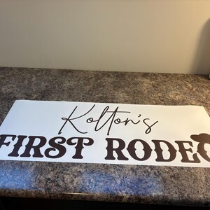 以下が含まれることがあります： Kolton's FIRST RODEOと黒い文字で書かれた白い長方形のビニールステッカー。Kolton'sは筆記体で、FIRST RODEOはカウボーイハットのアイコンが付いた太字のウェスタンスタイルのフォントで書かれています。
