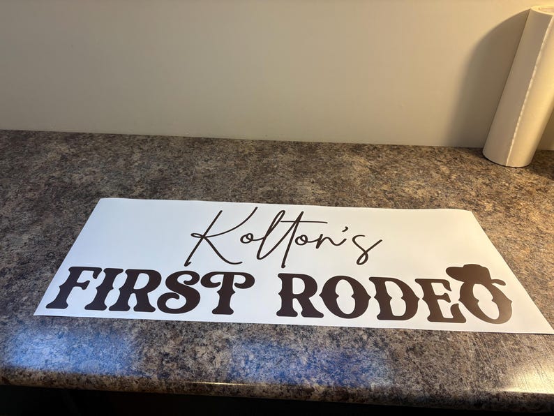 以下が含まれることがあります： Kolton's FIRST RODEOと黒い文字で書かれた白い長方形のビニールステッカー。Kolton'sは筆記体で、FIRST RODEOはカウボーイハットのアイコンが付いた太字のウェスタンスタイルのフォントで書かれています。