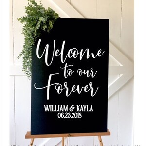 May include: A rectangular black sign featuring white lettering that reads Welcome to our Forever, followed by the names WILLIAM & KAYLA and the date 06.23.2018. A sprig of green eucalyptus leaves is attached to the top left corner of the sign.