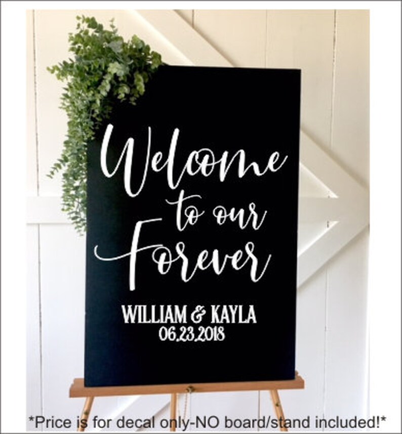 May include: A rectangular black sign featuring white lettering that reads Welcome to our Forever, followed by the names WILLIAM & KAYLA and the date 06.23.2018. A sprig of green eucalyptus leaves is attached to the top left corner of the sign.