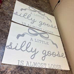 May include: Two white sheets of paper with the text "A little silly goose is almost loose" in gray script and block letters. The text is accompanied by gray ribbon-like illustrations. The sheets are placed on a brown countertop.