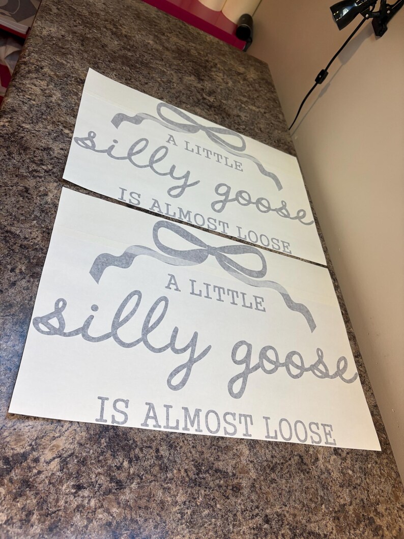 May include: Two white sheets of paper with the text "A little silly goose is almost loose" in gray script and block letters. The text is accompanied by gray ribbon-like illustrations. The sheets are placed on a brown countertop.