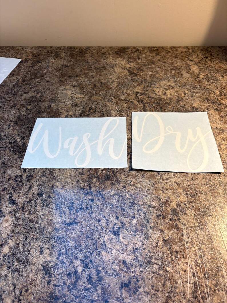 Puede incluir: Dos calcoman&iacute;as de vinilo blanco con texto en cursiva. Una dice "Wash" y la otra "Dry". Las calcoman&iacute;as est&aacute;n sobre una superficie marr&oacute;n moteada. Ideales para la decoraci&oacute;n del lavadero.