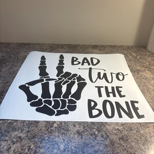 May include: A white vinyl decal featuring a black skeleton hand making a peace sign gesture. To the right of the hand, the text reads BAD two THE BONE in a combination of bold block lettering and a cursive script font for the word two.