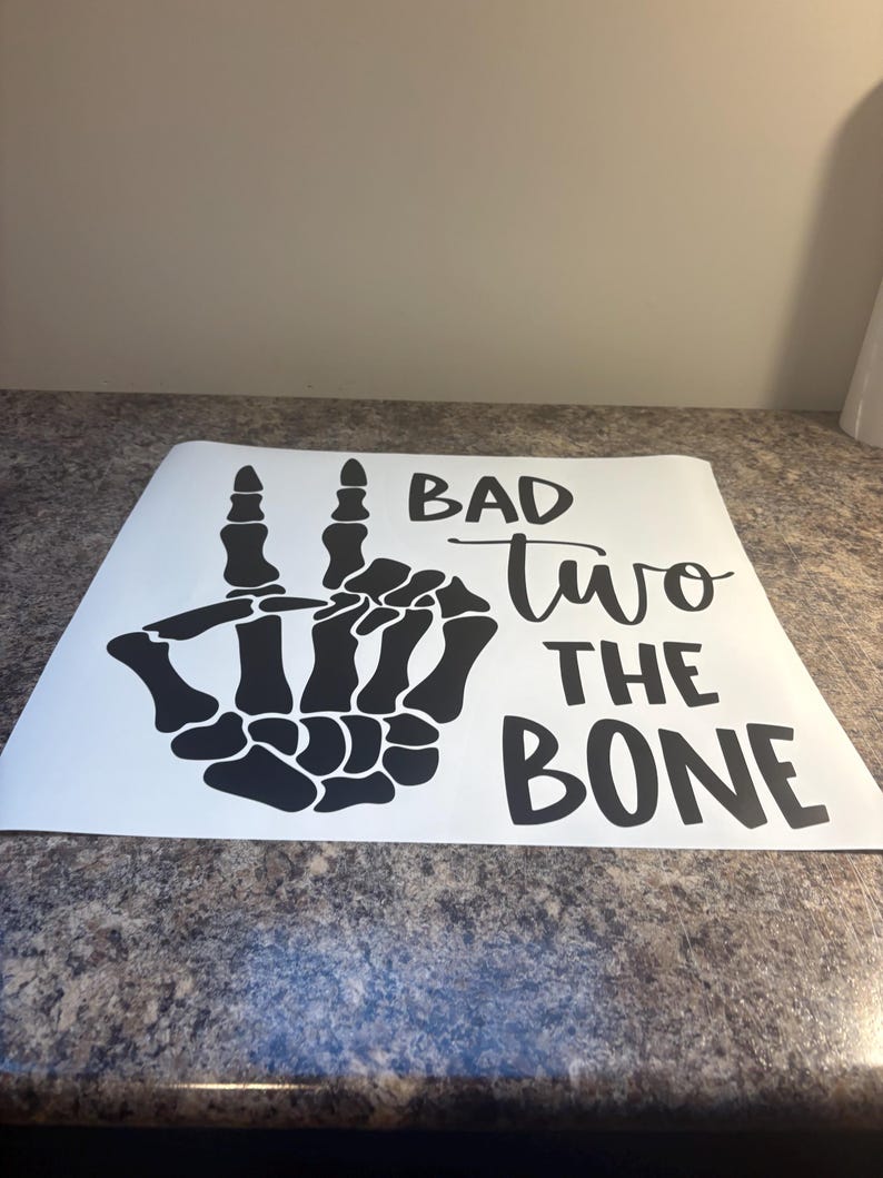May include: A white vinyl decal featuring a black skeleton hand making a peace sign gesture. To the right of the hand, the text reads BAD two THE BONE in a combination of bold block lettering and a cursive script font for the word two.