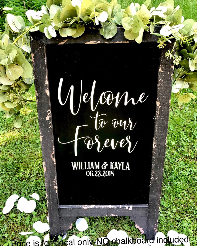 May include: A black chalkboard sign with a distressed wooden frame, decorated with green eucalyptus garlands. The sign features white text that reads Welcome to our Forever, WILLIAM & KAYLA, and the date 06.23.2018 in a mix of script and sans-serif fonts.