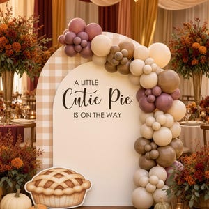 May include: An arched backdrop with a beige gingham pattern and the text A LITTLE Cutie Pie IS ON THE WAY. A balloon garland in shades of cream, tan, and mauve curves over the arch. A cutout of a pie sits at the base with assorted pumpkins.