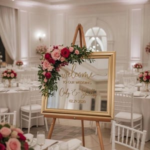 Może przedstawiać: Prostokątny lustrzany znak ślubny w złotej ramie na drewnianej sztaludze, z napisem "welcome TO OUR WEDDING Cody & Shelby 02.29.2020" złotą czcionką, ozdobiony bukietem czerwonych i r&oacute;żowych r&oacute;ż oraz zielonymi liśćmi w g&oacute;rnym rogu.