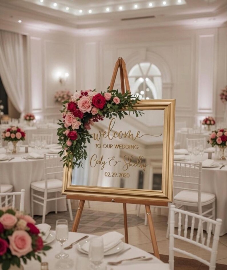Może przedstawiać: Prostokątny lustrzany znak ślubny w złotej ramie na drewnianej sztaludze, z napisem "welcome TO OUR WEDDING Cody & Shelby 02.29.2020" złotą czcionką, ozdobiony bukietem czerwonych i r&oacute;żowych r&oacute;ż oraz zielonymi liśćmi w g&oacute;rnym rogu.