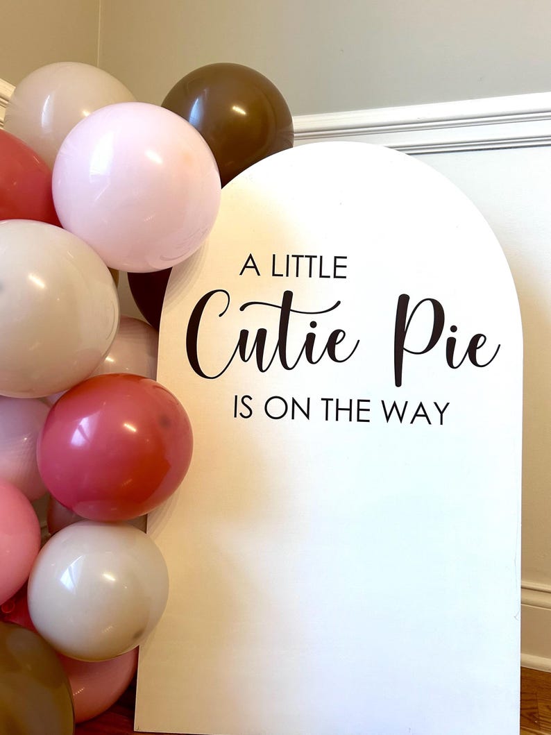 May include: A white arched sign with black text that reads A LITTLE Cutie Pie IS ON THE WAY, accompanied by a cluster of balloons in shades of pink, dark brown, and cream. The sign features a smooth surface and a rounded top edge.