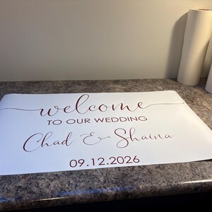 Może przedstawiać: Biała prostokątna naklejka winylowa z ciemnoczerwonym tekstem o treści welcome TO OUR WEDDING Chad & Shaina 09.12.2026. Projekt wykorzystuje połączenie eleganckiego pisma odręcznego dla imion i słowa welcome oraz prostych liter drukowanych.