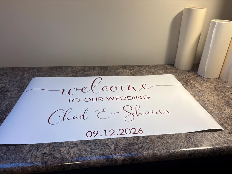 Może przedstawiać: Biała prostokątna naklejka winylowa z ciemnoczerwonym tekstem o treści welcome TO OUR WEDDING Chad & Shaina 09.12.2026. Projekt wykorzystuje połączenie eleganckiego pisma odręcznego dla imion i słowa welcome oraz prostych liter drukowanych.