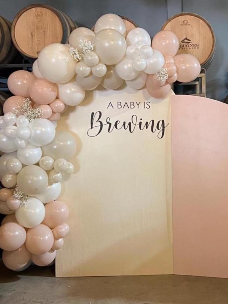 May include: A baby shower backdrop featuring a balloon arch in shades of white and blush pink. The arch frames a wooden panel with the phrase "A Baby Is Brewing" in elegant script. The backdrop is set against a backdrop of wooden barrels.