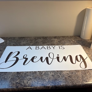May include: White sign with the phrase "A BABY IS Brewing" in a dark brown, elegant script. The sign is on a speckled countertop, with rolls of white material in the background.
