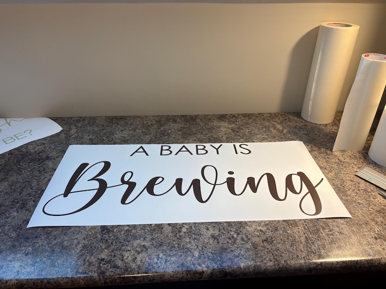 May include: White sign with the phrase "A BABY IS Brewing" in a dark brown, elegant script. The sign is on a speckled countertop, with rolls of white material in the background.