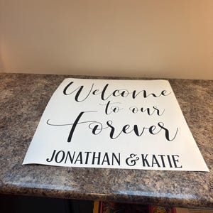 May include: A white rectangular sign featuring black text that reads Welcome to our Forever and JONATHAN & KATIE. The sign uses a combination of a decorative script font for the first three words and a bold sans-serif font for the names.
